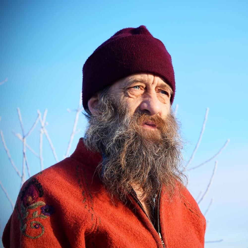 A bearded man with a maroon beanie looks into the distance outside.